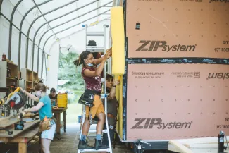 Students work on a tiny house in an outdoor classroom. A student with braids uses a nail gun on the tiny house siding in the foreground.