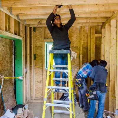 A student standing on ladder running electrical wire in a ceiling cavity.