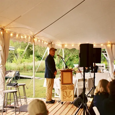 John Connell stands at a podium under a white celebration tent addressing a seated crowd. He is wearing a baseball cap and suit jacket.
