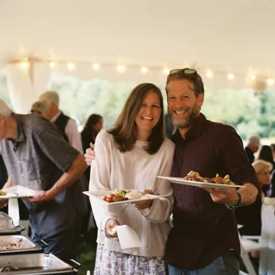 Sarah and Giles, holding plates of food, smile at the camera under a white tent