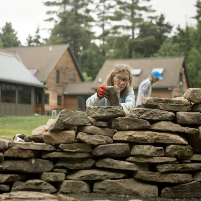 A student stacking stone in a partially complete stone wall with other students in the background.