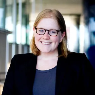Abby Martin smiles at the camera in front of a blurred office scene. She has short, blonde hair, and is wearing glasses, a black blazer, and blue shirt.