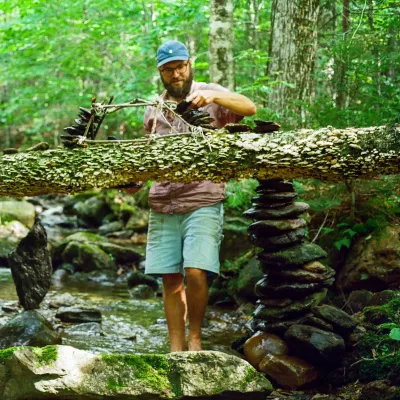 Adam stands in a shallow brook in a forest, building a small structure on a fallen tree. He is wearing shorts, red shirt, and a hat.