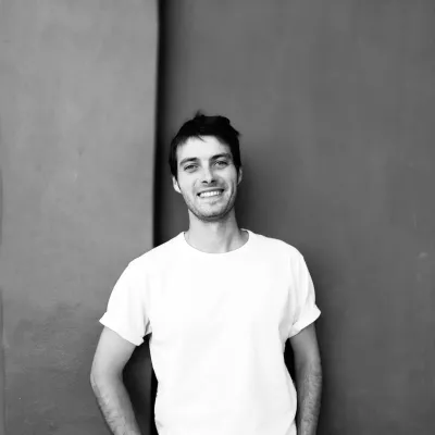 Alex stands in front of a wall smiling at the camera. The photo is black and white, he has his hands in his pockets and is wearing a white t-shirt.