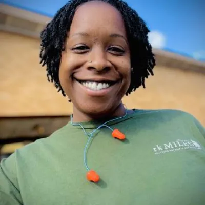 Anastasia Gladmon stands in front of a building with sky in the background. She has short black locs, and is wearing a green t-shirt with earplugs around her neck.