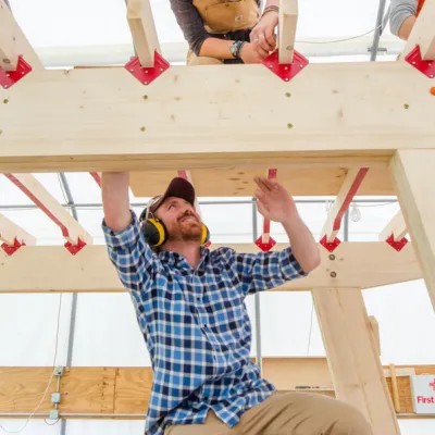 Andy White stands on a ladder looking up while building the roof of a structure.