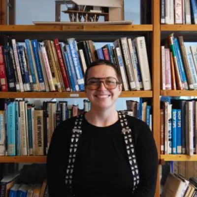 Anna Fluri, standing in front of bookshelves, smiles at the camera. She has short, brown hair, glasses, and is wearing a black shirt and suspenders.