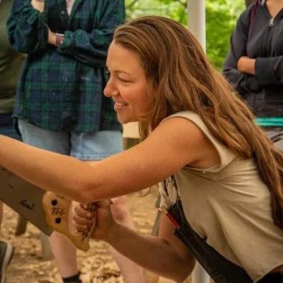 Ariel is kneeling down and cutting joinery on a beam. She has long, light brown hair and is wearing a white t-shirt.