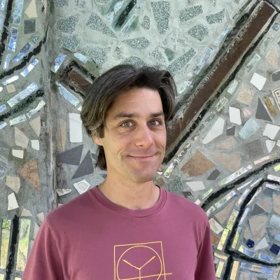 Aron smiles at the camera in front of a tile mosaic wall. He has medium-length dark hair and is wearing a raspberry colored yestermorrow t-shirt.
