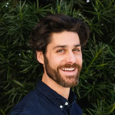 Aron smiles at the camera in a headshot with trees in the background