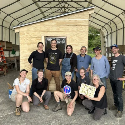 A group of twelve people poses for a photo in front of their garden shed.
