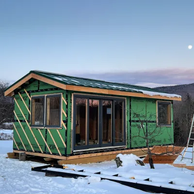 A zip-sheathed cabin sits on a snowy hillside at dusk with the moon and ridgeline in the background. 