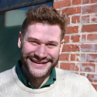 Ben smiles at the camera in front of a brick wall. He is wearing a white sweater and has short, light brown hair and beard.