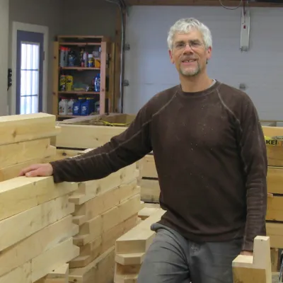 Ben stands with his hand on a pile of lumber next to him. He is wearing a green sweater and glasses.