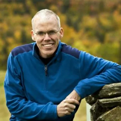Bill McKibben stands outside, leaning against a rock wall with foliage in the background. He is wearing glasses and a blue fleece sweatshirt.