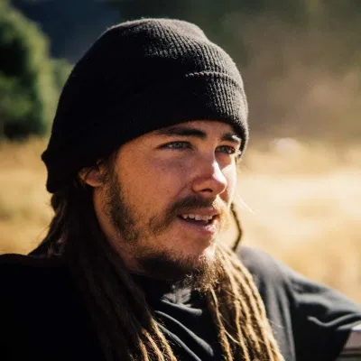 Brenden is photographed outside, looking to the right of the camera. He is wearing a dark beanie and has long brown hair.