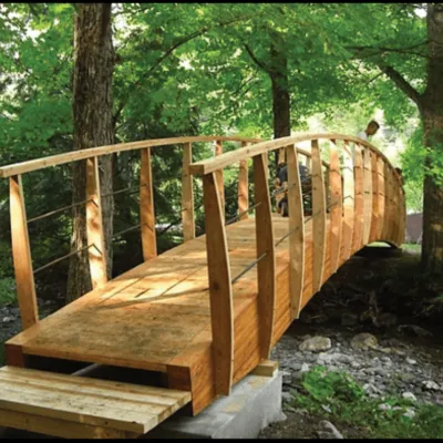A wooden bridge spans a shallow brook in the forest. The are two adults working on the bridge.
