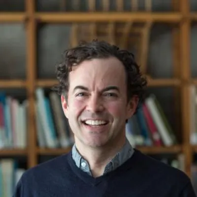 Britton Rogers, standing in front of books, smiles at the camera. He has short, dark curly hair and a blue sweater.