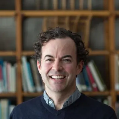 Britton Rogers, standing in front of books, smiles at the camera. He has short, dark curly hair and a blue sweater.
