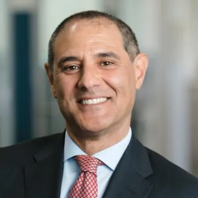 Bruno Grimaldi smiles at the camera in front of a blurred background. He has short grey hair and is wearing a black suit with a red tie.