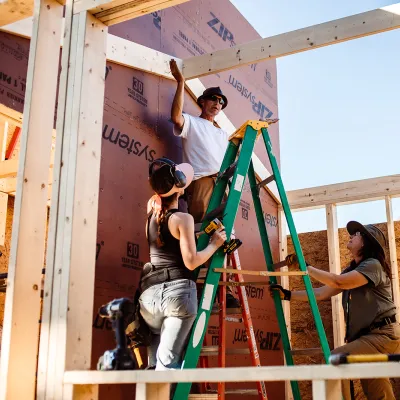 An instructor on a ladder gestures at a partially framed building. 