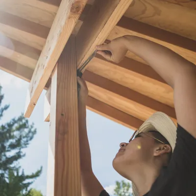 A person uses an angle finder on the corner of an outdoor structure, with trees and sky in the background. The photo is shot from below, looking up at the person reaching up to the corner. 