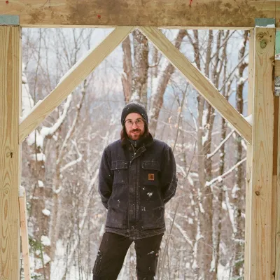 Charlie stands outside in winter under an unfinished door frame. He is wearing black work clothes and has his hands behind his back.