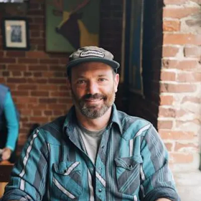 Chris Lucchesi sitting at a table smiling at the camera. He is wearing a brown cap and blue flannel shirt. He has short, brown facial hair.