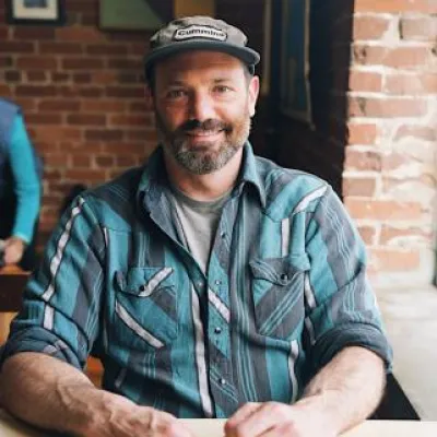 Chris Lucchesi sitting at a table smiling at the camera. He is wearing a brown cap and blue flannel shirt. He has short, brown facial hair.