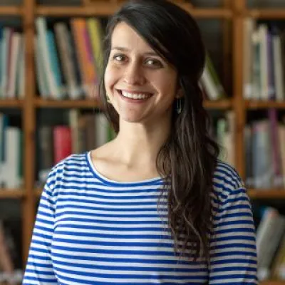 Chrissy Bellmyer, standing in front of bookshelves, smiles at the camera. She has long, dark brown hair and is wearing a blue and white striped shirt.