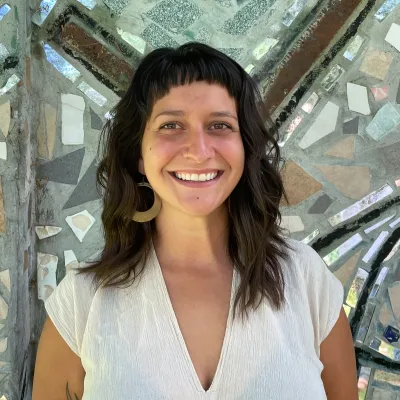 Chrissy is standing in front of a mirrored mosaic and smiling at the camera. She had shoulder length brown hair and bangs. She's wearing a white shirt and hoop earrings. 
