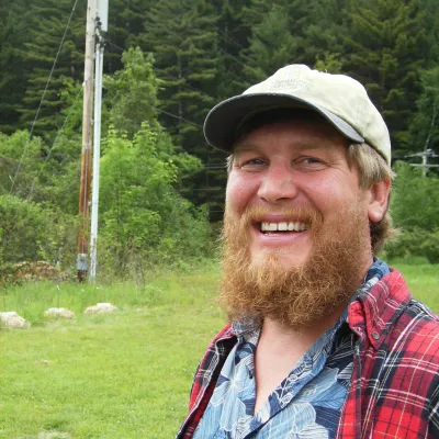 Christian smiles at the camera, standing outside with trees and grass in the background. He is wearing a red flannel shirt and baseball cap. He has a long light brown beard and blue eyes.