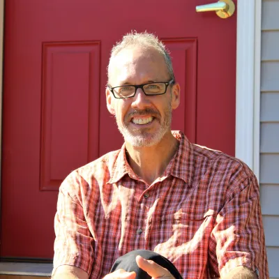 Cullen Ryan smiles at the camera outside in front of a red door. He is wearing glasses and a red plaid collared shirt.