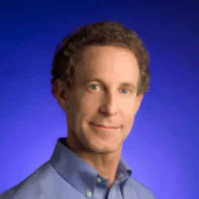 Dan Reicher smiles at the camera in front of a solid blue background. He is wearing a blue collared shirt and has short, curly brown hair.