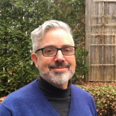 Danny looks at the camera, smiling. He has a royal blue turtleneck sweater and glasses.