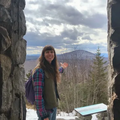 Dawn stands outside with mountains in the background. She has long brown hair and straight bangs and is wearing a backpack.