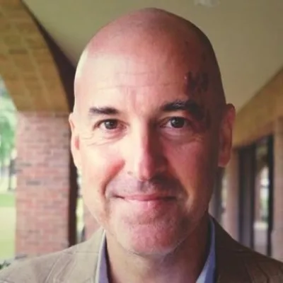 Dodge Bingham smiles at the camera, outside in front of a house. He is bald with brown eyes and wearing a beige blazer.