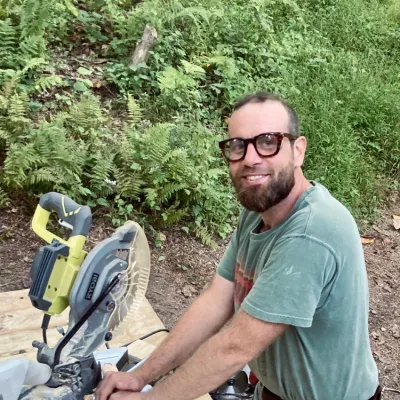Drew smiles at the camera at a chop saw station. He is wearing a green t-shirt and eye protection.