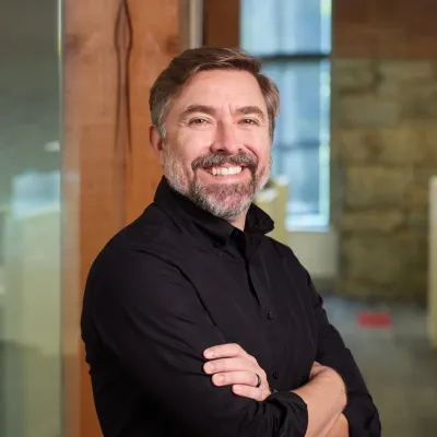 Drew smiles at the camera with his arms crossed. He has short dark hair, facial hair, and is wearing a black collared shirt.