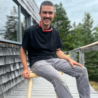 Eli sits on a wooden chair, outside on the deck of a grey shingled building. He has short dark hair, black t-shirt, and grey pants.