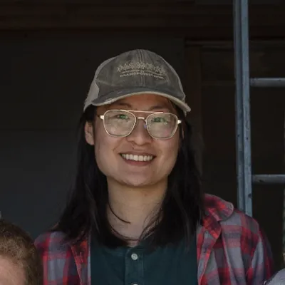 Emily smiles at the camera. She has shoulder length dark hear and is wear a baseball cap, glasses and a flannel. 