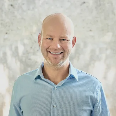 Erik smiles at the camera in front of a white background. He is wearing a light blue button-down shirt.