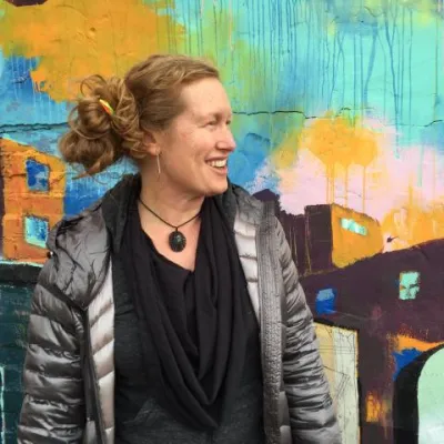Erin stands in front of a colorful mural, looking to the right and smiling. She has curly red hair pulled back into a bun and is wearing a silver puffy jacket.