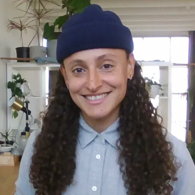Evie smiles at the camera inside. She has long, dark brown curly hair and is wearing a blue beanie and blue collared shirt.