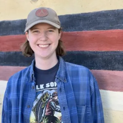 Ezrie Anderson stands in front of a beige, blue, and red wall. She is smiling, wearing a beige baseball hat and blue flannel shirt.
