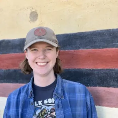 Ezrie Anderson stands in front of a beige, blue, and red wall. She is smiling, wearing a beige baseball hat and blue flannel shirt.