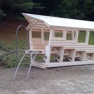 A two-story wooden playground structure with a metal roof and arched climbing bars sits on a gravel site against a backdrop of trees.