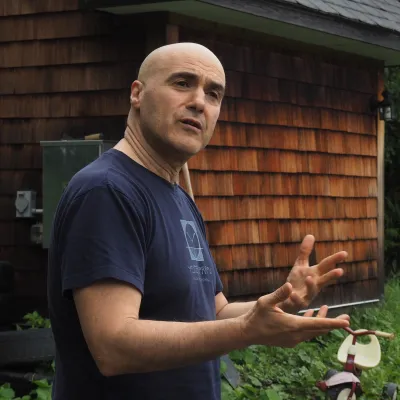 Gio is standing outside of a shingled building, gesturing with his hands. He is wearing a dark blue Yestermorrow t-shirt.