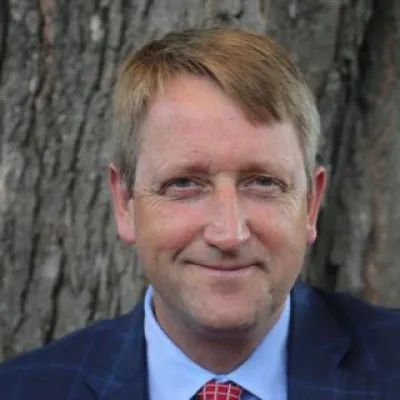 Gunnar Hubbard stands in front of a tree, smiling at the camera. He has short, light hair and is wearing a blue suit with a red tie.