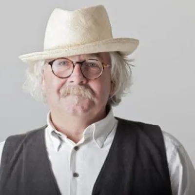Hank Louis stands in front of a white background, smiling at the camera. He is wearing a white brimmed hat, a white button-down shirt and black vest.